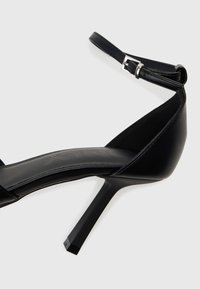 Svart högklackad sandal med en elegant design, som har ett glansigt läderövre, en tunn ankelsnodd och en fyrkantig klack.