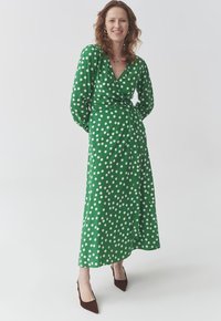 Langes grünes Wickelkleid mit weißen Punkten, langen Ärmeln und einer gebundenen Taille. Kombiniert mit braunen spitzen High Heels.