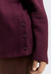 Pull marron avec un design de tricot texturé, comportant quatre boutons sombres sur le côté et une coupe décontractée. Tissu doux et manches longues.