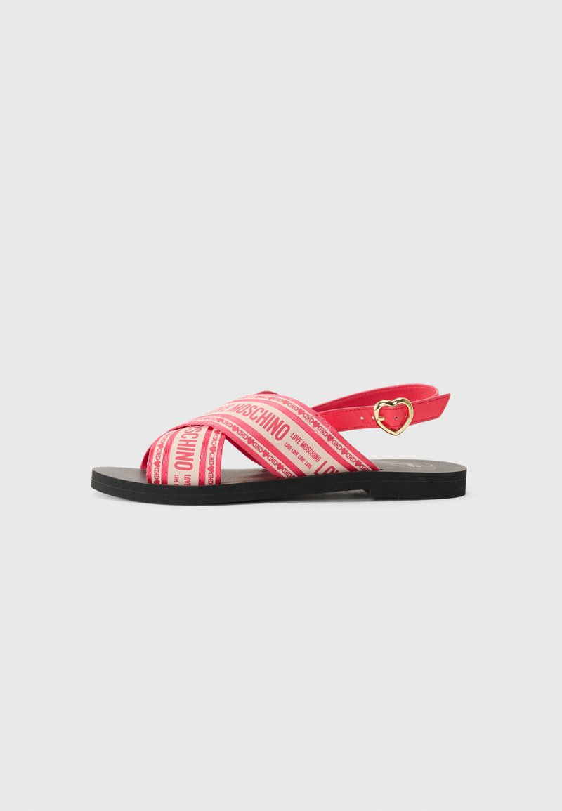 Red sandals with cross-strap design made from synthetic material, featuring printed logo and heart-shaped hardware accent. Flat black sole.