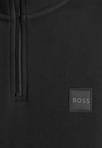 Gros plan sur un tissu noir montrant une fermeture éclair verticale et une pièce carrée cousue avec le mot « BOSS » en lettres noires assorties.