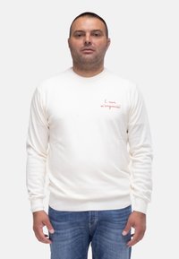 Maglione crema con maniche lunghe e scollatura rotonda. Presenta testo ricamato rosso sul petto. Indossato con jeans blu.