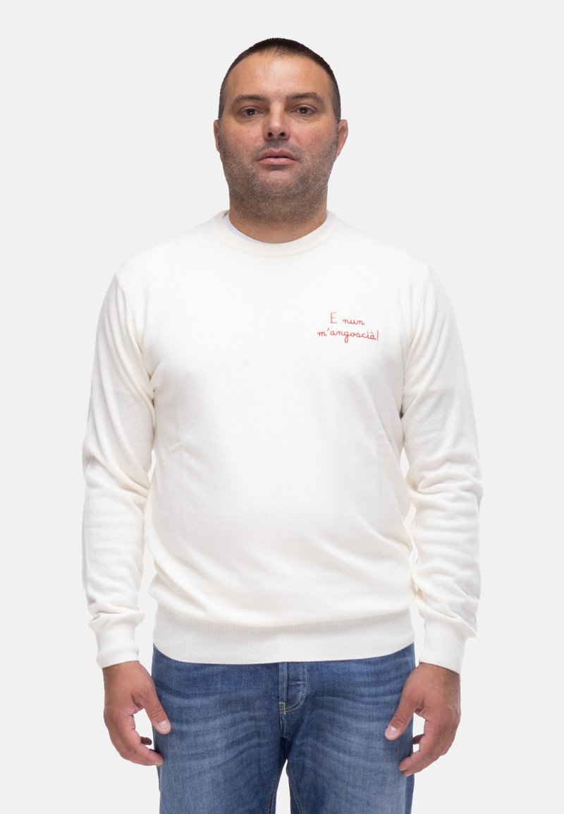 Maglione crema con maniche lunghe e scollatura rotonda. Presenta testo ricamato rosso sul petto. Indossato con jeans blu.