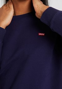 Tmavě modrá mikina z měkké bavlny, s malým červeným logem Levi's na hrudi a žebrovanými manžetami. Svetr má volný střih a kulatý výstřih.