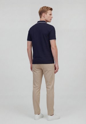 Mann trägt ein dunkelblaues Poloshirt mit weißem Rand, beigefarbene Hose und weiße Turnschuhe, steht mit dem Rücken zur Kamera vor einem schlichten grauen Hintergrund.