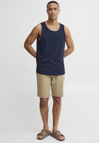 Zeleno-modra bombažna majica brez rokavov z žepom, v kombinaciji z bež kratkimi hlačami. Model nosi rjave sandale, kar prikazuje minimalističen dizajn in sproščen stil.