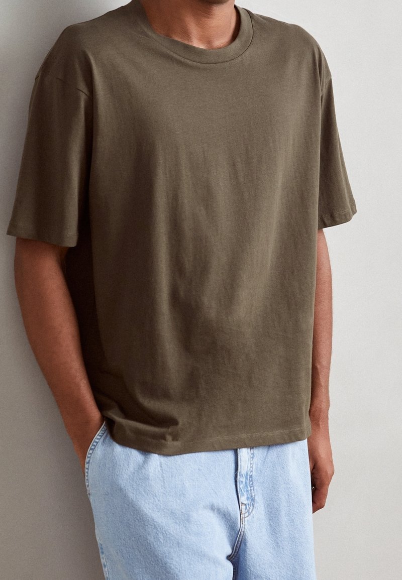 Person wearing an olive green oversized short-sleeve t-shirt and light blue denim jeans with hands in pockets against a plain wall.