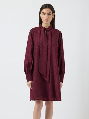 Bordeauxfarbenes Kleid aus strukturiertem Stoff, mit langen Ärmeln, Bindeband am Hals und lockerem Schnitt, das bis zur Mitte des Oberschenkels reicht.