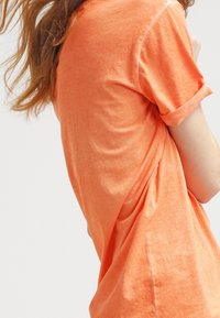 Osoba v jasně oranžovém tričku s krátkým rukávem a ohrnutými manžetami, s vlasy splývajícími, mírně otočená od kamery na bílém pozadí.