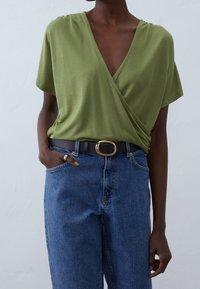 Olive green short-sleeved wrap top with a deep V-neck, paired with high-waisted blue denim jeans and a black belt with a gold buckle.