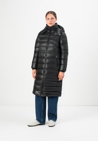 Vrouw in een lange, zwarte gewatteerde puffer jas met een staande kraag en rits, gecombineerd met blauwe jeans en witte schoenen.