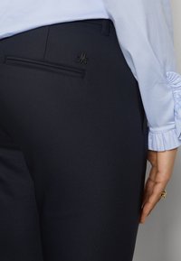 Gros plan d'une personne portant un pantalon noir avec un détail de poche arrière et un chemisier bleu clair avec des poignets froncés sur la manche.