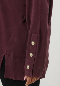 Ärm av en vinröd stickad tröja med tre dekorativa guldknappar nära handleden, bärs över svarta byxor, hand delvis synlig.