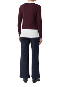 Person in einem burgunderfarbenen Pullover über einem weißen Hemd, mit dunklen weiten Hosen und schwarzen Schuhen, steht mit dem Rücken zur Kamera.