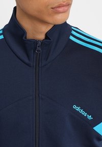 Marinblå zip-up jacka med ståkrage, har blå ränder längs axlarna och en broderad Adidas-logotyp på bröstet. Slät textur.