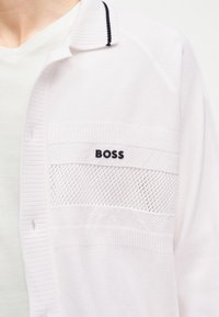 Λευκή πλεκτή ζακέτα με διάτρητο πάνελ και μαύρο λογότυπο "BOSS" στο στήθος, φορεμένη πάνω από λευκό T-shirt.