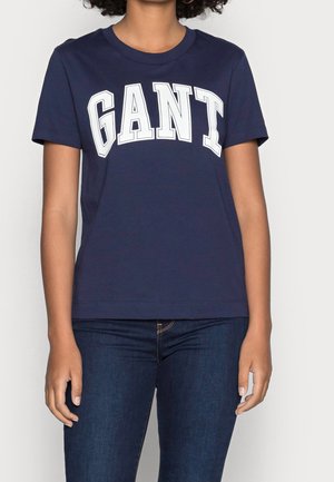 Osoba nosząca granatowy T-shirt z dużym białym tekstem "GANT" i ciemnoniebieskie dżinsy, stojąca na jednolitym jasnym tle.