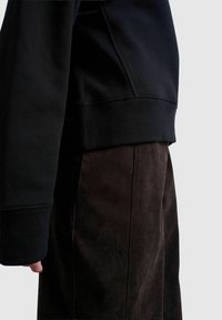 Vue de profil d'une personne portant un sweat-shirt noir avec des poignets côtelés et un pantalon en daim brun foncé sur un fond uni.