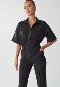 Zwarte jumpsuit met korte mouwen, een ritssluiting, twee voorzakken en een taillering voor een getailleerde pasvorm. Soepele, lichtgewicht stof.