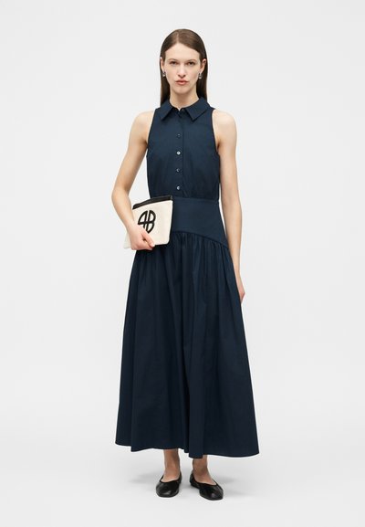 Žena stojaca v tmavomodrých maxi šatách bez rukávov so zapínaním na gombíky, drží bielu kabelku s čiernym logom a má na nohách čierne balerínky.