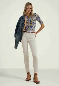 Kvinne iført blomstermønstret topp, hvite bukser, brun belte og platåsandaler, som holder en denimjakke over skulderen mot en ensfarget bakgrunn.