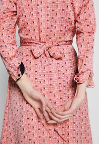 Femme portant une robe rose à motifs avec manches longues et ceinture nouée à la taille, mains doucement jointes dans le dos, sur un fond uni.