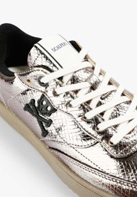 Zapatillas plateadas metálicas con acabado texturizado, cordones blancos, detalle de logo en cuero negro y suela de goma color marrón claro.