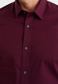 Burgundy button-up skjorta med krage, med en slät yta och mörka knappar, synliga sömmar och en figurnära design.