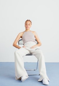 Šedý halter top s tenkými ramínky, doplněný volnými bílými tepláky s logem, kulatými bílými teniskami, sedící na židli.