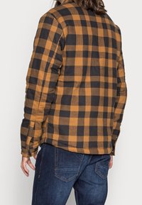 Brun och svart rutmönstrad skjortjacka med vadderad textur, med knäppning och sidofickor, som bärs tillsammans med mörka denimjeans.