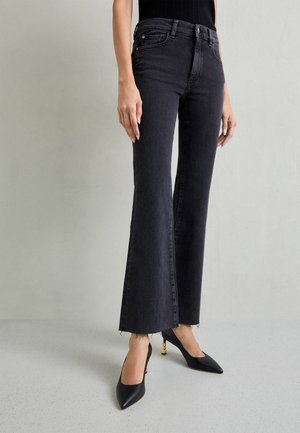 Kvinde iført sorte højttaljede flare jeans med rå kanter og sorte spidse hæle med gulddetalje på hælen.