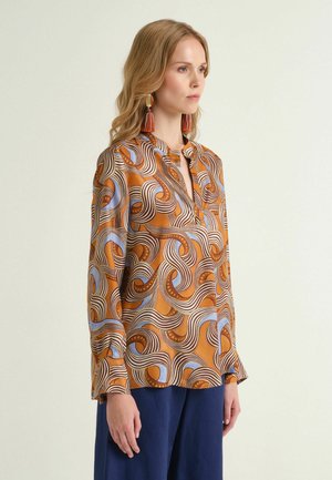 Femme portant un chemisier à manches longues aux motifs orange, bleu et marron, avec un pantalon bleu marine et des boucles d'oreilles à pompons orange, vue de profil.