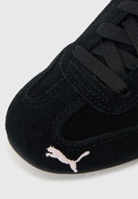 Μαύρα σουέτ αθλητικά παπούτσια με υφή, που διαθέτουν μαύρες κορδόνια και λευκό λογότυπο Puma. Είναι ορατό το στρογγυλό εμπρός μέρος και οι λεπτομέρειες ραφής.