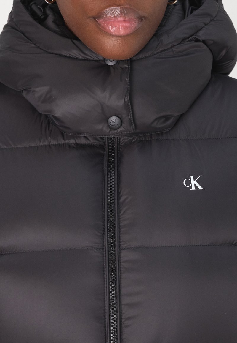 Schwarze Steppweste mit hohem Kragen, Frontreißverschluss und Druckknöpfen. Verfügt über ein dezentes Logo auf der Brust und eine glatte, glänzende Stoffoberfläche.