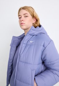 Svetlo vijoličasta napihnjena jakna s kapuco, ki ima diskreten logotip Nike, prešito vzorčenje, zadrgo in stranske žepe. Gladka tekstura.