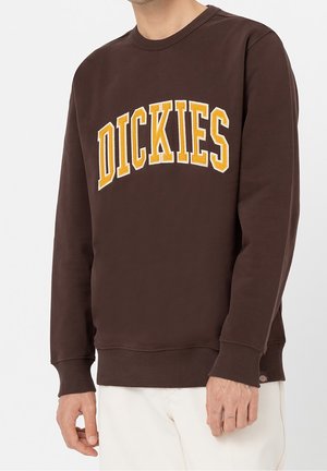 Mężczyzna ubrany w brązową bluzę z żółtym napisem "DICKIES" na piersi oraz kremowe spodnie, na jednolitym tle.