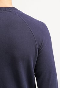 Gros plan de l'arrière d'une personne portant une chemise à manches longues bleu foncé avec des coutures visibles le long de l'épaule.