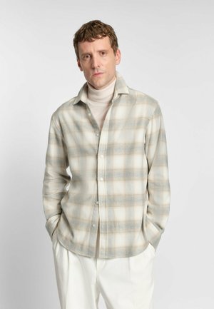Camicia a quadri beige con chiusura a bottoni, maniche lunghe e texture morbida. Presenta accenti di blu chiaro e marrone. Indossata sopra un dolcevita chiaro.
