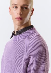 Pull en maille lavande avec texture côtelée et encolure ronde. Porté par-dessus une chemise à carreaux foncés, présentant un motif de points en relief sur les manches.