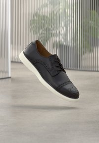 Chaussure à lacets en cuir noir avec un bout perforé et des détails décoratifs le long des côtés, avec une semelle en caoutchouc blanche.