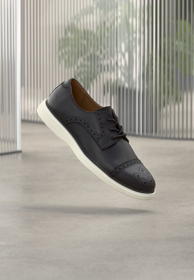 Chaussure à lacets en cuir noir avec un bout perforé et des détails décoratifs le long des côtés, avec une semelle en caoutchouc blanche.