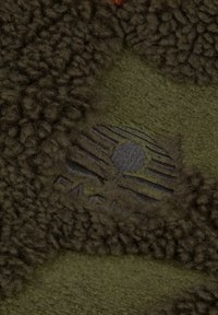 Närbild av brun texturerad fleece med ett broderat grön och grå Timberland-logotypmärke.