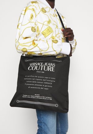 Černá taška typu tote s bílým textem "VERSACE JEANS COUTURE", vyrobená z látky, obdélníkového tvaru a s pevnou rukojetí.