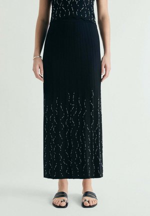 Mujer con falda larga negra acanalada con patrones ondulados recortados, combinada con sandalias negras abiertas con detalles de hebilla plateada.