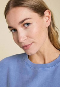 Sweat-shirt bleu clair avec un col arrondi et des détails de couture, porté par une personne à la peau claire et aux cheveux blonds raides.