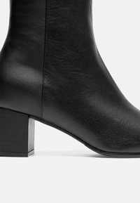 Bottine en cuir noir avec un talon carré, texture lisse et détails de couture minimalistes sur le côté.