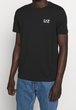 T-shirt noir en coton à manches courtes, col rond, avec un logo EA7 blanc sur la poitrine gauche ; associé à un jean en denim bleu.