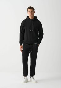Sudadera negra con cordones ajustables, combinada con pantalones jogger negros con dobladillos ajustados y acento de logo. Usada con zapatillas blancas.