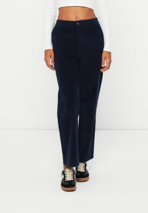 Femme portant un pantalon droit taille haute bleu marine foncé, un top blanc court à manches longues et des baskets noires avec des lacets blancs.