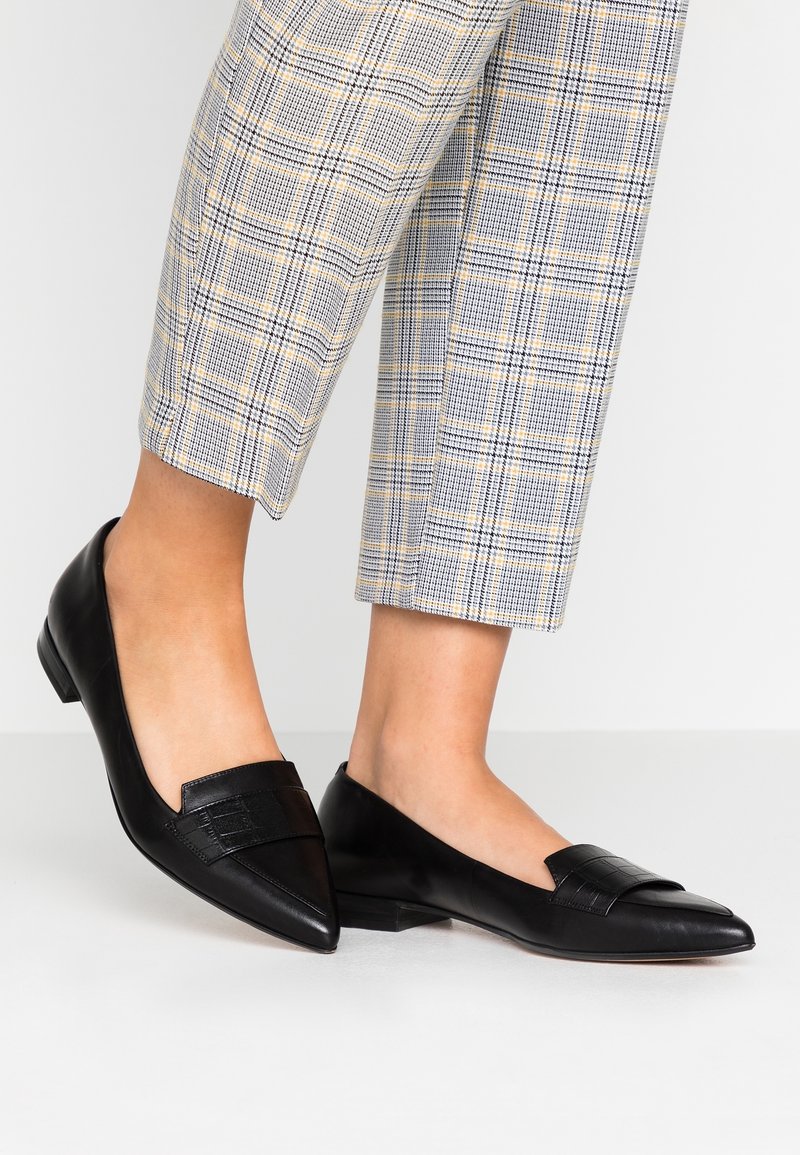 Jambes portant un pantalon écossais coupé et des mocassins noirs à bout pointu, debout sur un sol clair devant un fond blanc.